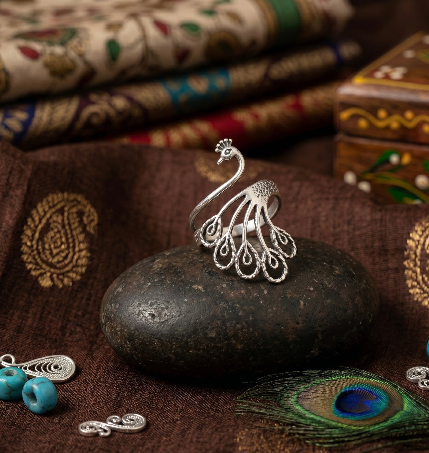Silver peacock-shaped ring on a dark stone with decorative fabric in the background
