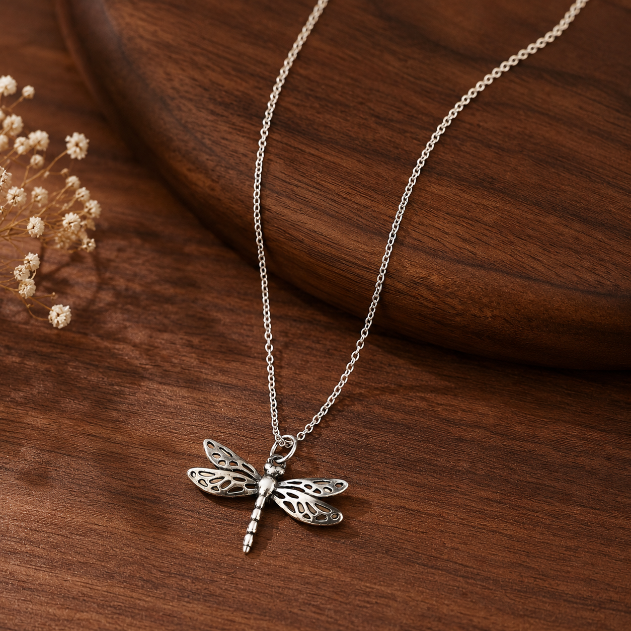 A silver dragonfly pendant necklace on a delicate chain, displayed on a dark brown wooden surface with dried flowers in the background.