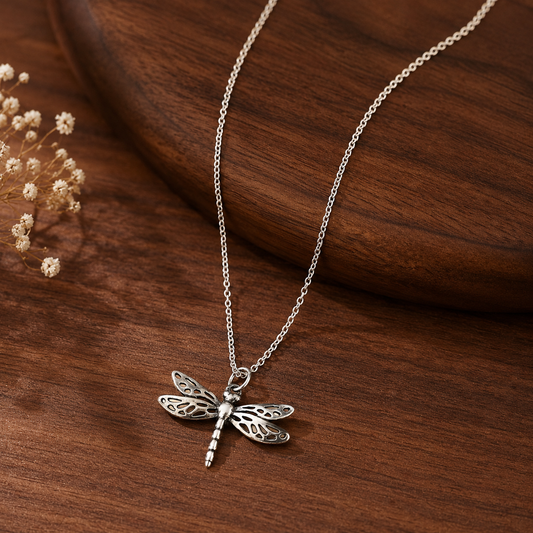 A silver dragonfly pendant necklace on a delicate chain, displayed on a dark brown wooden surface with dried flowers in the background.
