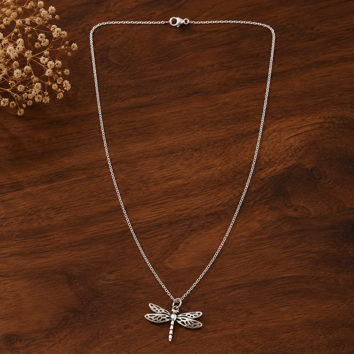 A silver dragonfly pendant necklace on a delicate silver chain, displayed on a dark brown wooden surface with dried flowers in the top-left corner.