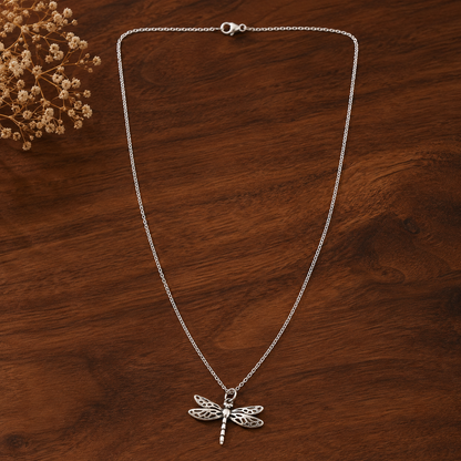 A silver dragonfly pendant necklace on a delicate silver chain, displayed on a dark brown wooden surface with dried flowers in the top-left corner.