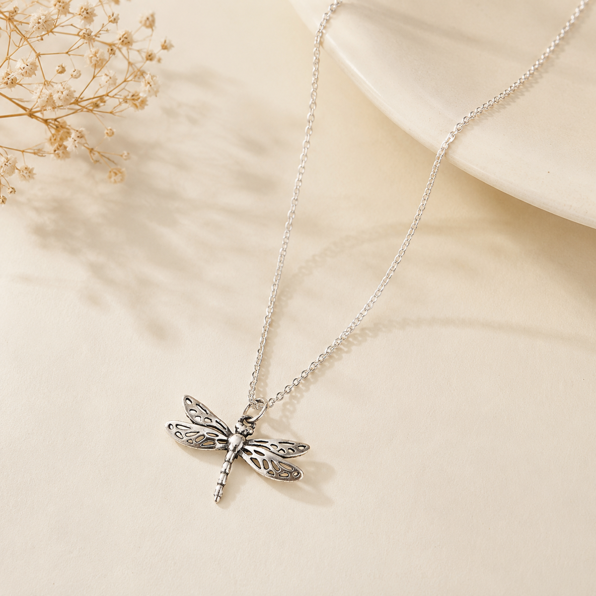 A silver dragonfly pendant necklace on a delicate silver chain, styled on a warm beige surface . Made in pure 925 sterling by silveristhan