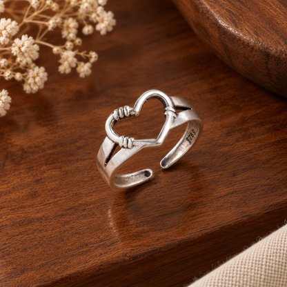 A sterling silver open ring with a heart-shaped top and textured wrap details, displayed on a polished dark brown wooden surface with dried flowers in the background.