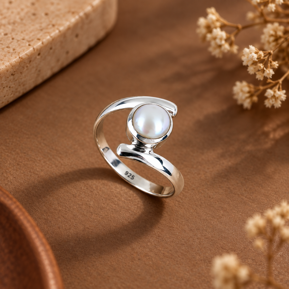 A sterling silver ring with a round white pearl, featuring a polished 925 hallmark on the inner band, styled on a textured brown surface with dried flowers.