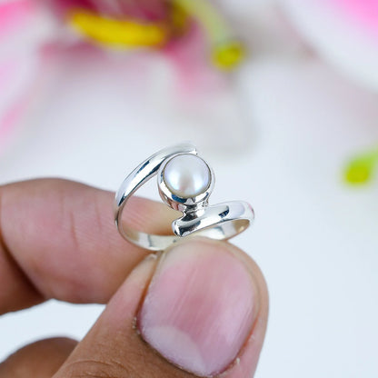 A silver ring with a round white pearl, held between two fingers against a soft, blurred pink and white background.
