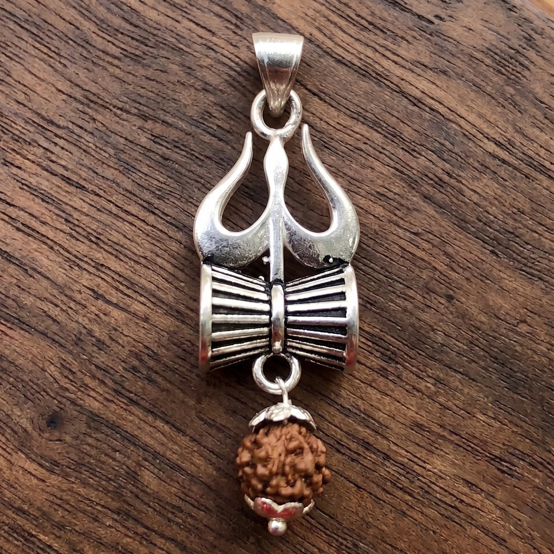 Close Up image of Silver Trishul Damru pendant with hanging panchmukhi rudraksh on a dark wooden surface by silveristhan
