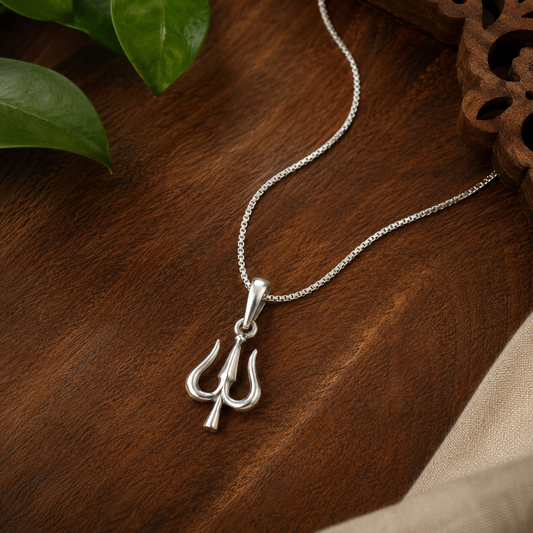 A silver trident pendant necklace on a matching chain, displayed on a dark wooden surface with green leaves and a beige fabric prop.