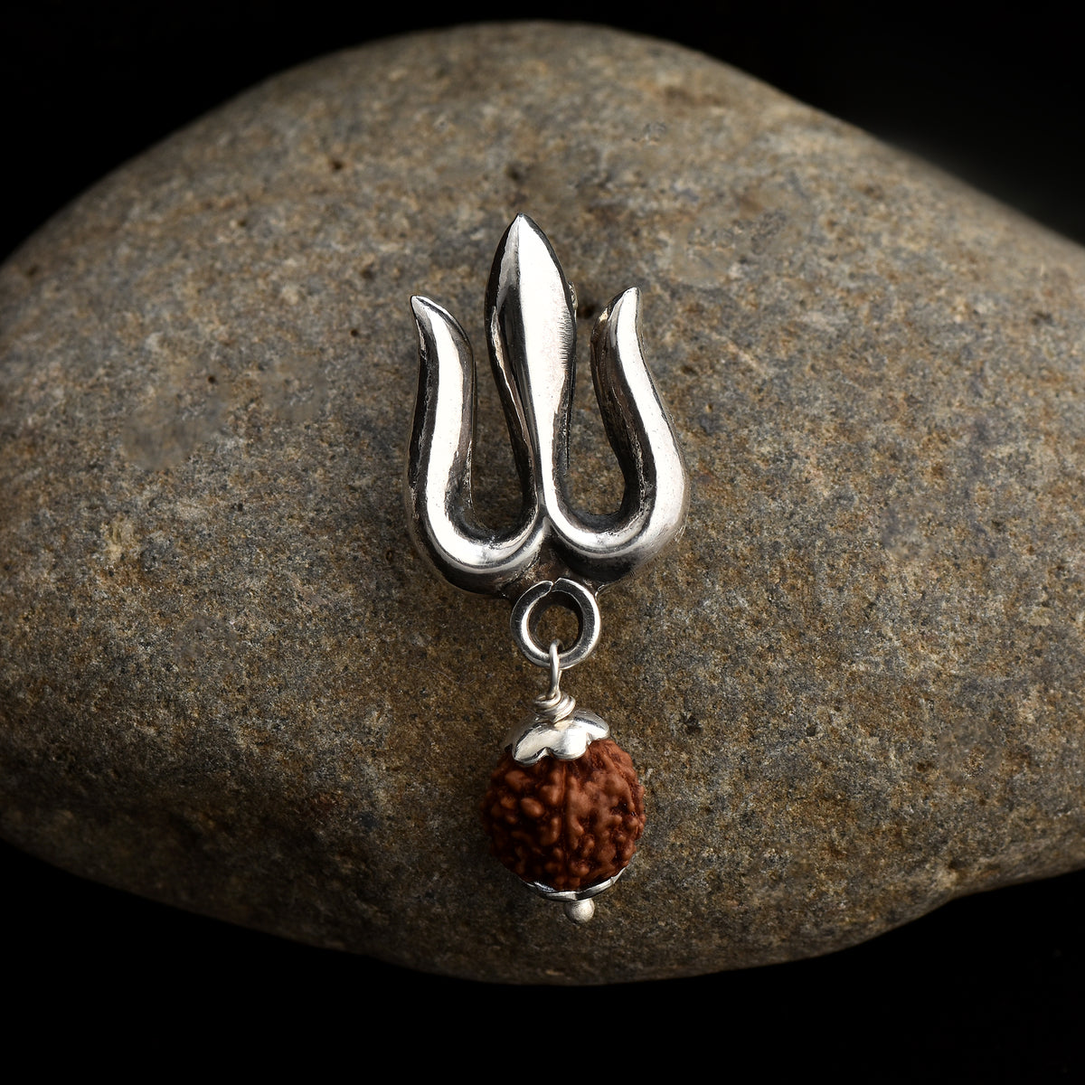 A silver trident-shaped brooch with a sacred Rudraksh bead attached at the bottom, displayed on a rock.