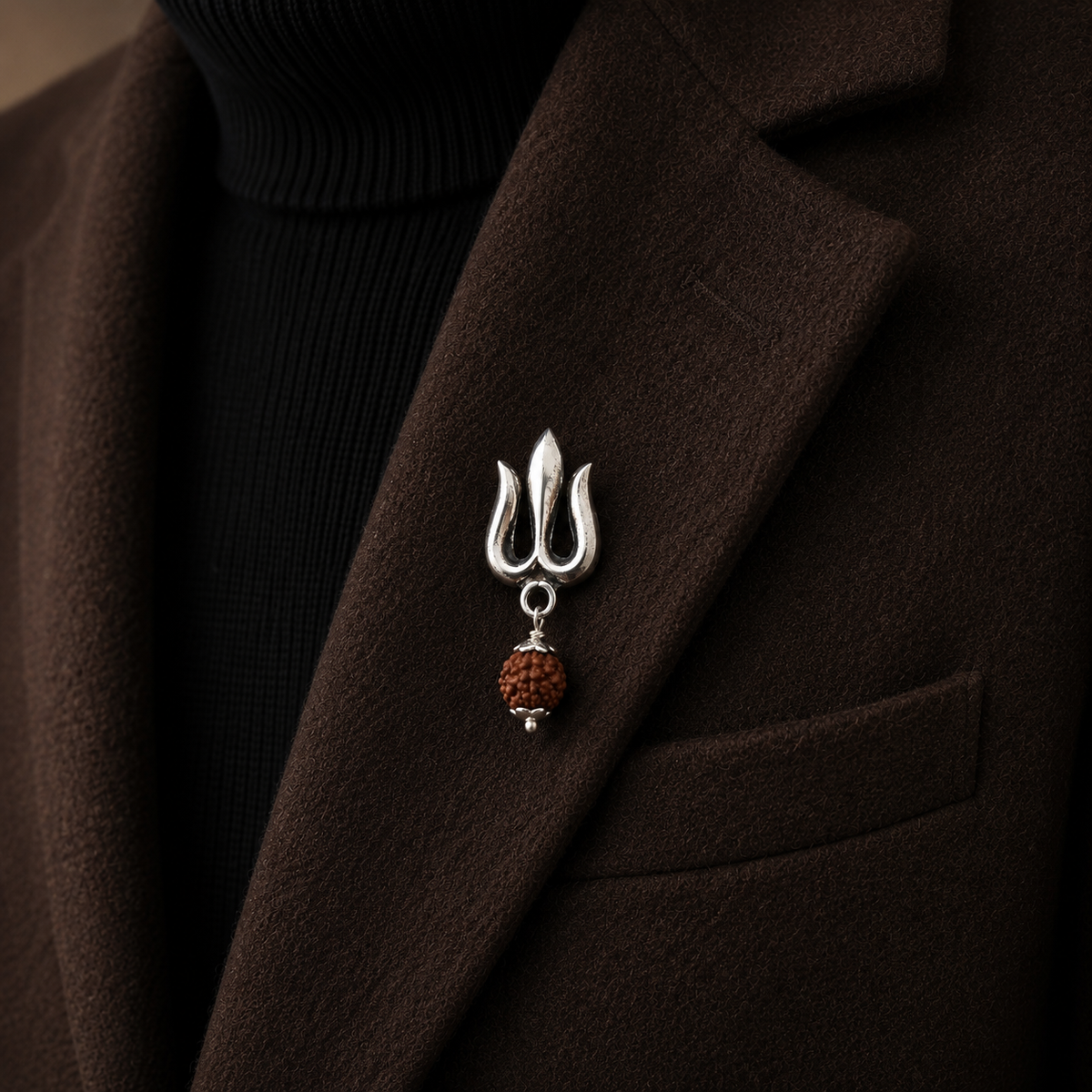 A sterling silver made brooch shaped like a trident with hanging rudraksh, pinned to the lapel of a dark brown coat over a black ribbed turtleneck.