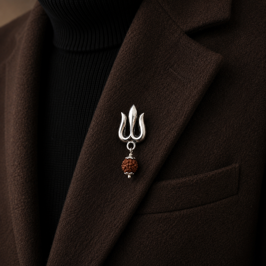 A sterling silver made brooch shaped like a trident with hanging rudraksh, pinned to the lapel of a dark brown coat over a black ribbed turtleneck.