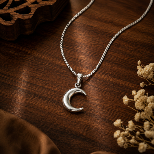 A silver crescent moon pendant necklace on a delicate silver chain, displayed on a dark wooden surface with dried flowers and a brown fabric accent.